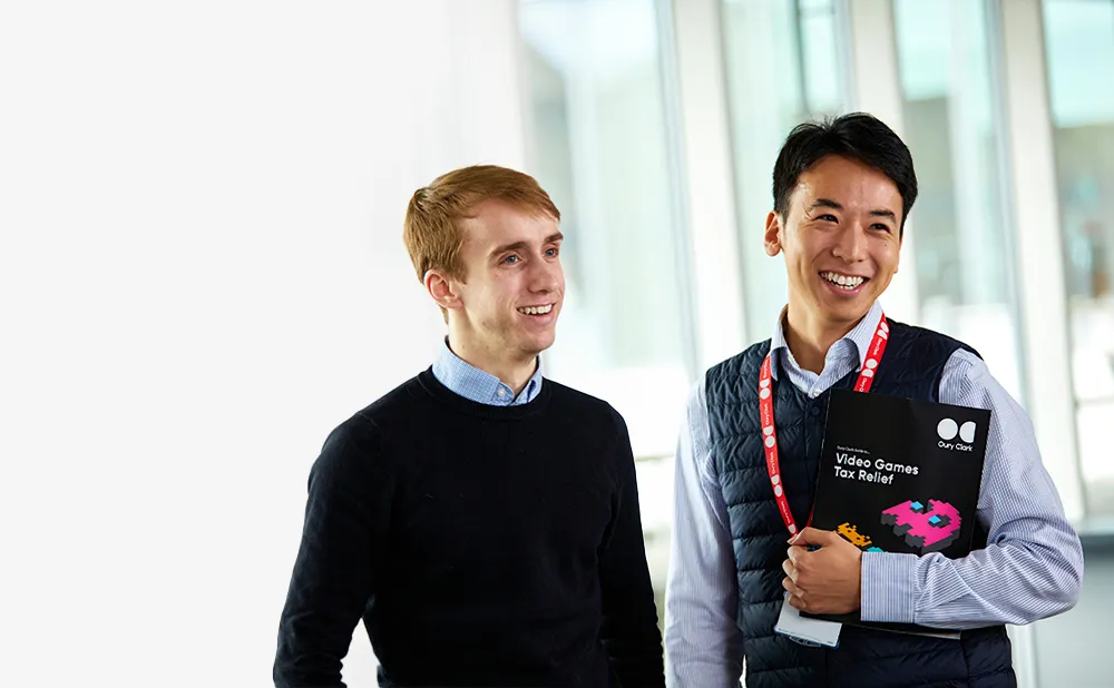 Photo of two smiling men, the one on the right is weating a lanyard and holding a copy of an Oury Clark guide to video games tax relief